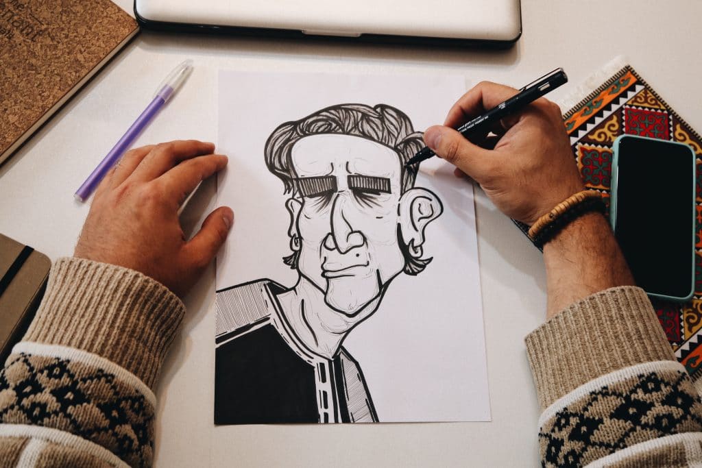 A man's hands are visible drawing a characterization of a face in black pen. 