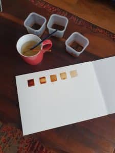 A cup with a paintbrush in it, and three plastic containers with coffee inside them, a piece of wtarecolor paper with 5 squares painted with coffee. This shows tonal variation examples when painting with coffee.