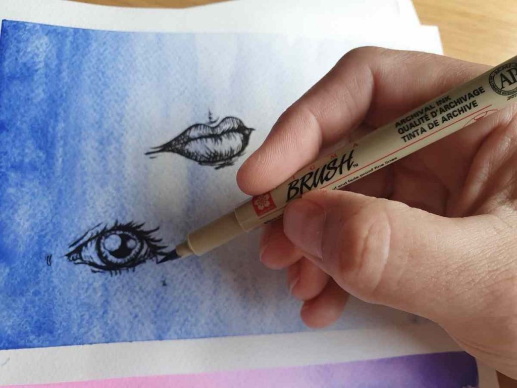 a hand demonstrating how to use micron pens on a watercolor background